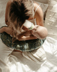 A mother sits on a bed feeding her baby with a bottle, using a DockATot nursing pillow for support. She's wearing a green patterned skirt and a white top, with sunlight filtering through creating a warm atmosphere.