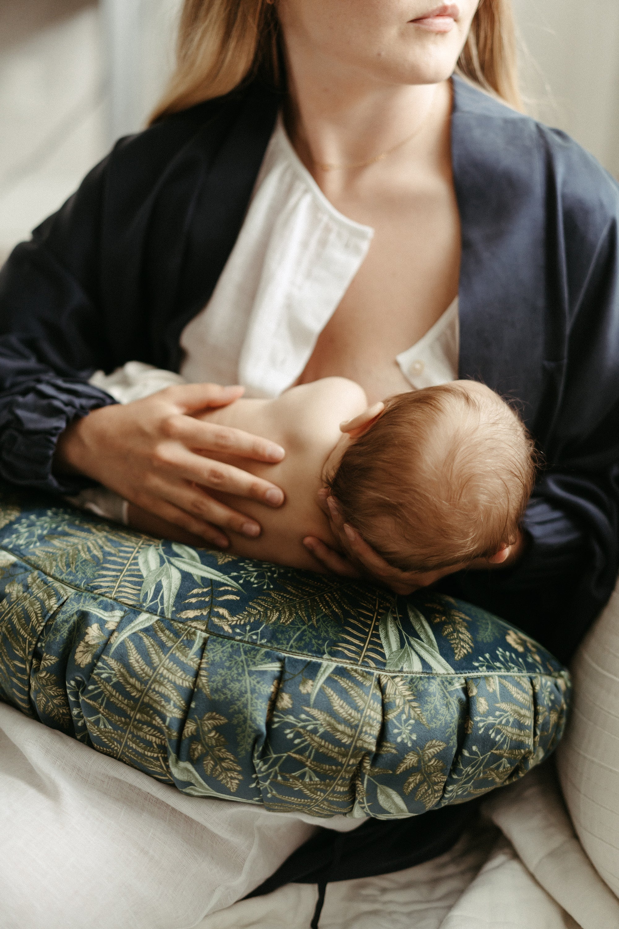 A mother in a white shirt and blue robe breastfeeding her baby, who is supported by a DockATot La Maman Wedge - Navy Night Falls, in a soft-lit room. Only the back of the