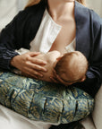 A mother in a white shirt and blue robe breastfeeding her baby, who is supported by a DockATot La Maman Wedge - Navy Night Falls, in a soft-lit room. Only the back of the