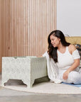 A woman sits on a round beige rug in a bright room, looking at an open book next to a DockATot Kind Essential Bassinet - Willow Boughs with ornately crafted pedestal feet. Wooden panels and a plant.