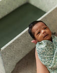 A newborn baby wrapped in a floral blanket looks towards the camera, held by a caregiver whose face is partially visible. They are seated near a DockATot Kind Essential Bassinet - Willow Boughs with ornately crafted pedestal feet in a softly.