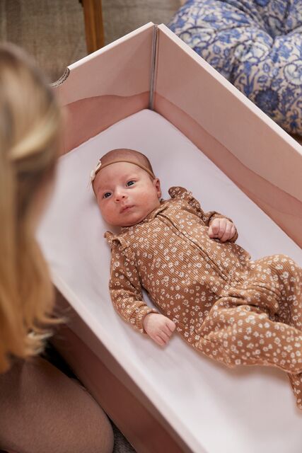 A newborn baby lies in a pink DockATot Kind Essential Bassinet with a white sheet, dressed in a brown onesie with a floral pattern, gazing upward. A woman, visible from the back and partially blurred.